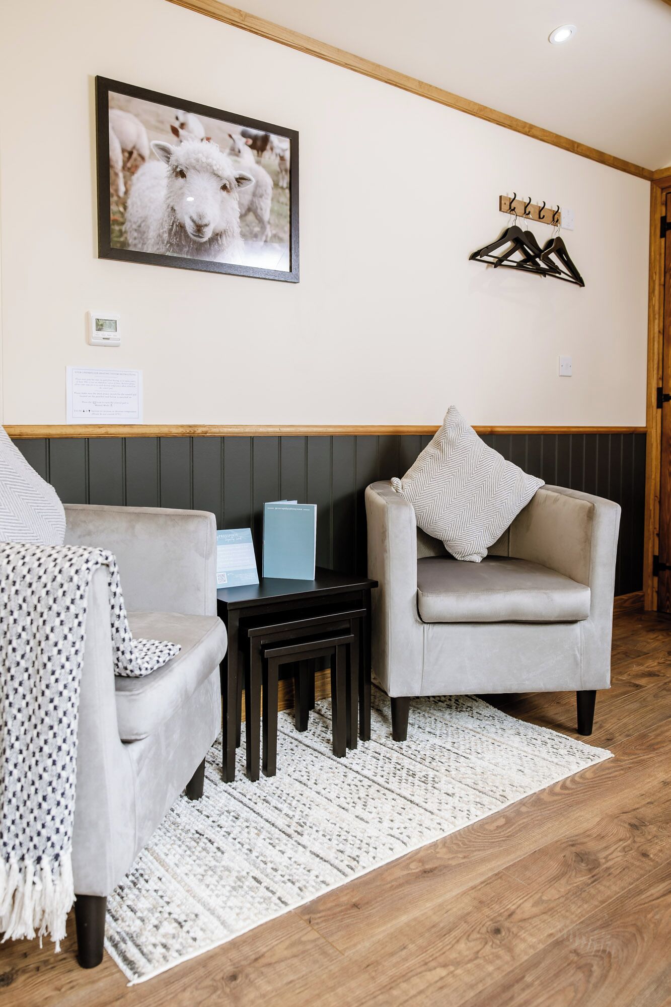 A cozy room with two grey armchairs, a small table, a framed picture of sheep, a coat rack, and a thermostat.