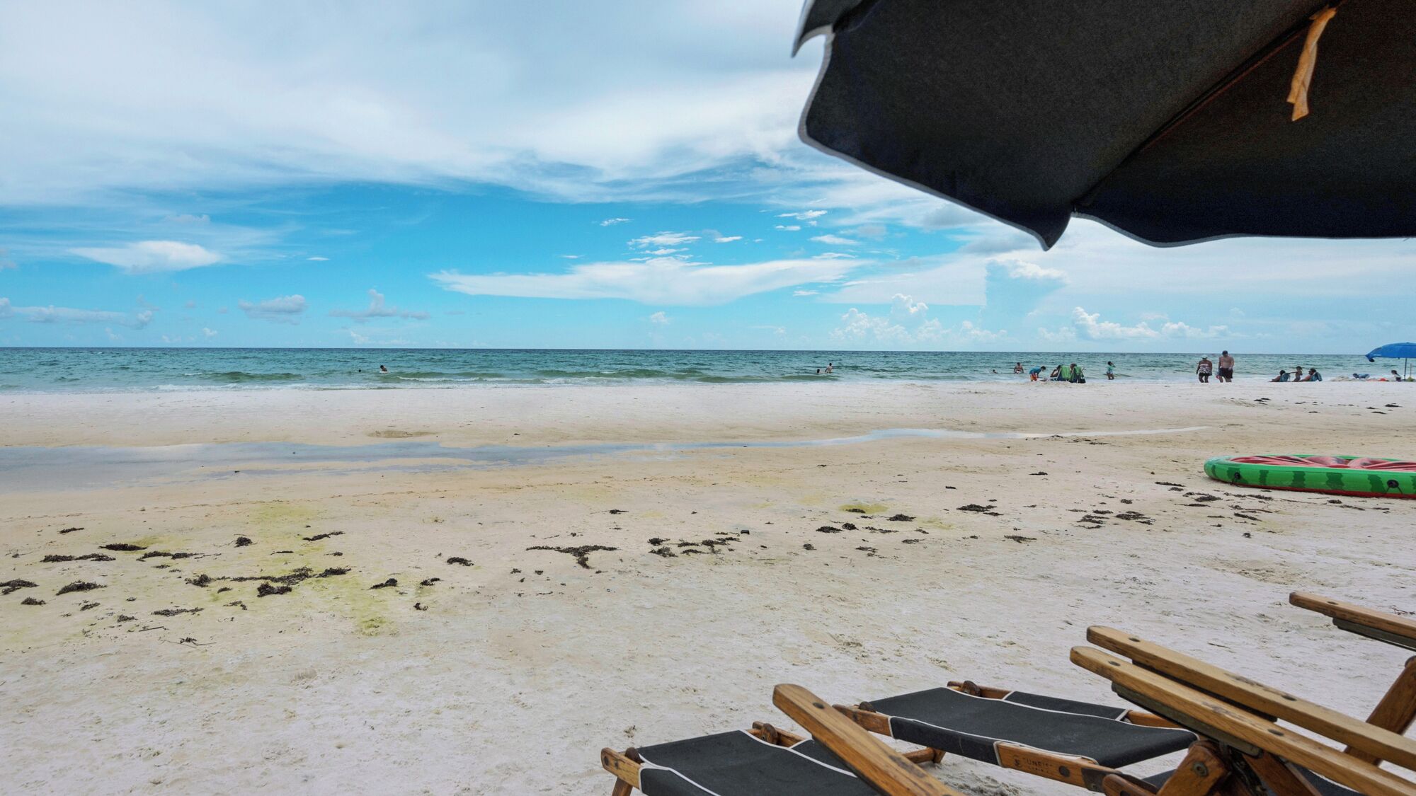 Une plage avec un ciel dégagé, un rivage sablonneux et quelques personnes dans l’eau.