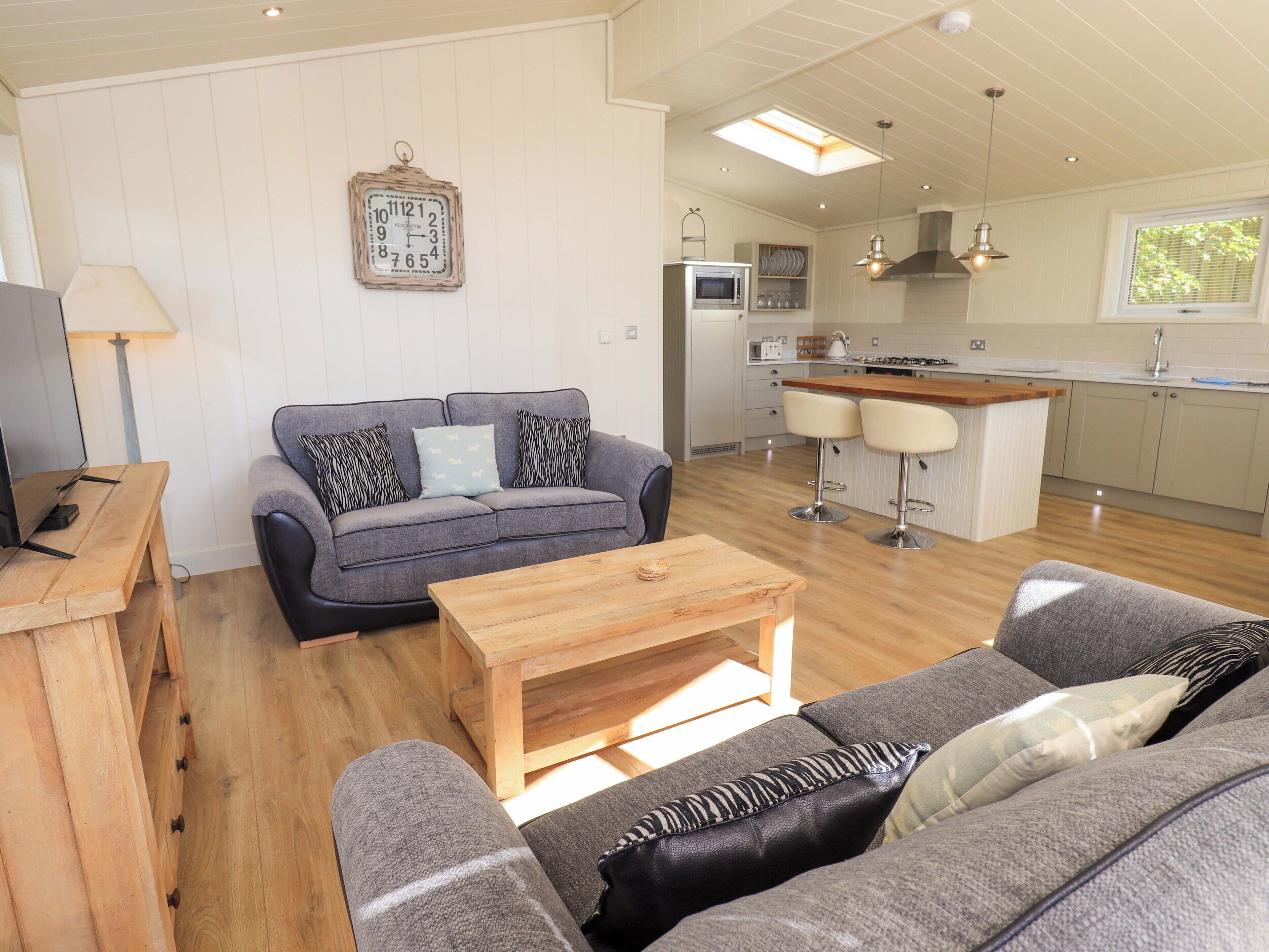 A modern living room with a grey sofa, wooden coffee table, and a TV on a wooden stand. A kitchen with a central island and bar stools is visible in the background.