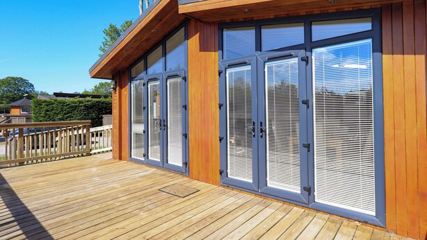 A modern building with large sliding glass doors, a wooden deck, and a clear blue sky. - Kingfisher Lodge (Skipton)