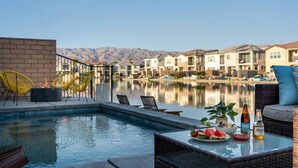 A poolside area with chairs, a table with food and drinks, and a view of houses across a body of water. - Solmar at Terra Lago Private Lake House With Free Pool Heating for any 3nights stay (Indio)