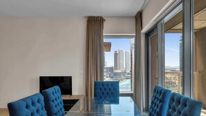 A modern dining area with a glass table and blue upholstered chairs, a flat-screen TV, and a view of a cityscape through large windows. - Bellavista - Elegant -2BR -Fountain View (Dubai)