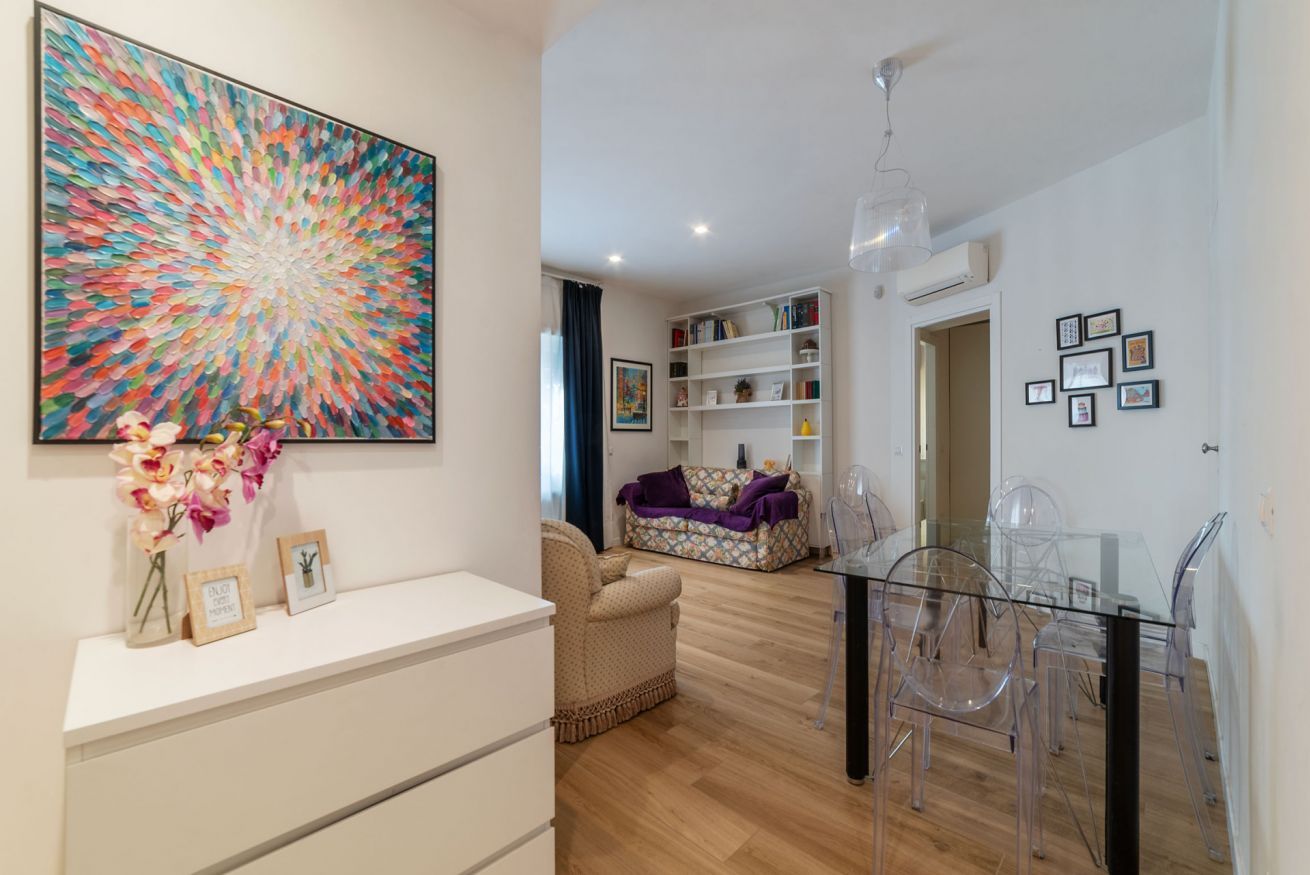 A modern living room with a glass dining table, clear chairs, a sofa, a bookshelf, and a large abstract painting.
