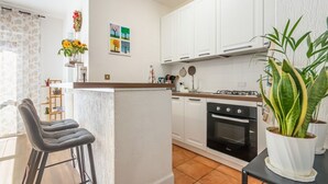 Eine Küche mit weißen Schränken, einer zentralen Kochinsel, einem Herd und einem Backofen. Auf der Insel stehen zwei Barhocker und ein Blumentopf.