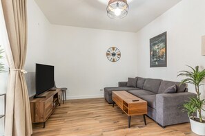 A modern living room with a grey sofa, a wooden coffee table, a TV on a wooden stand, a wall clock, and a painting.
