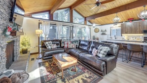 A cozy living room with a fireplace, leather sofa, and a large window overlooking a kitchen. - Modern Lake Arrowhead Retreat: 3 Mi to Village (Lake Arrowhead)