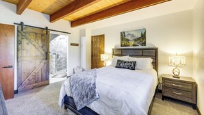 A bedroom with a large bed, wooden ceiling beams, a sliding barn door, a painting, and a nightstand with a lamp. - Modern Lake Arrowhead Retreat: 3 Mi to Village (Lake Arrowhead)