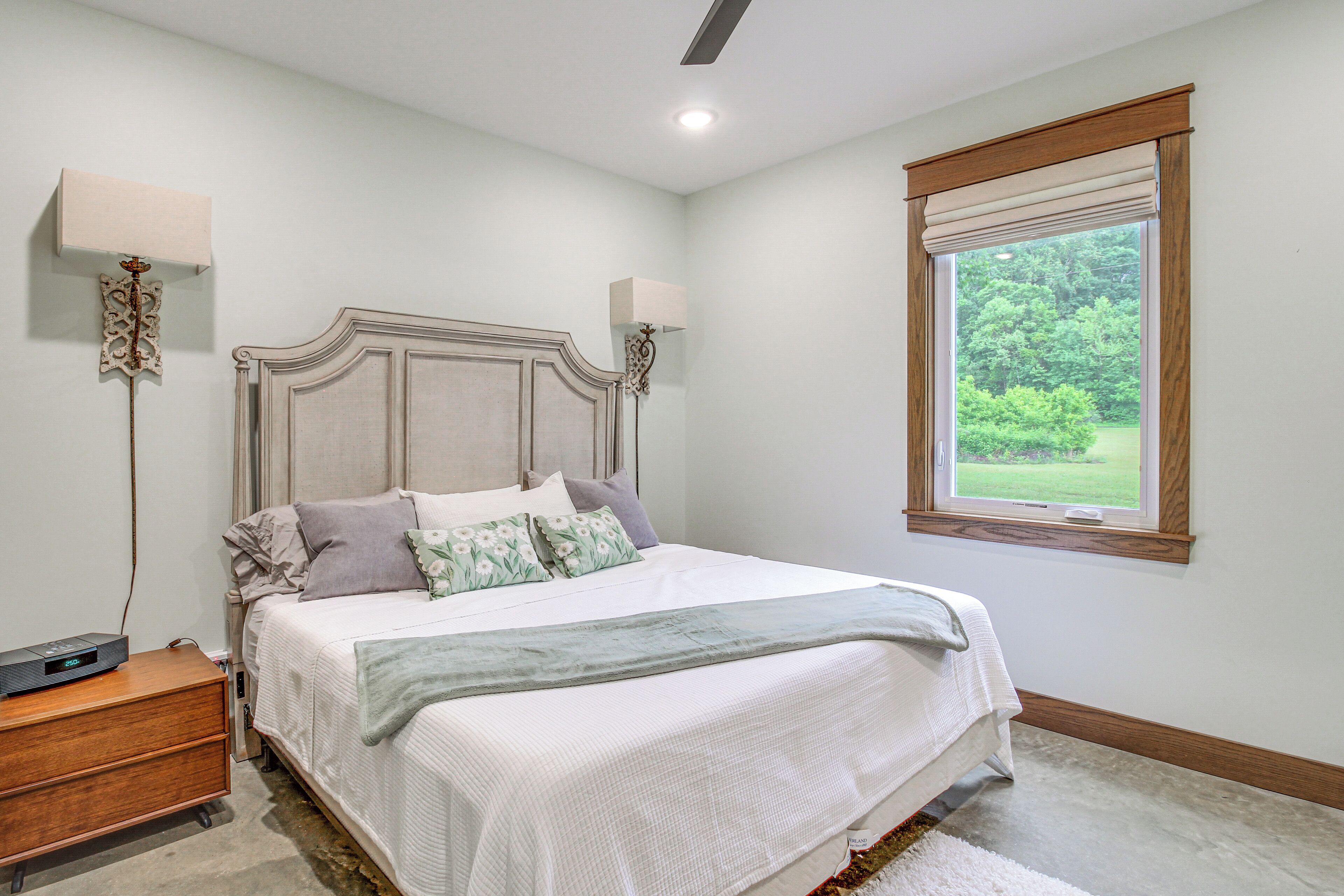 A bedroom with a large bed, bedside lamps, a nightstand, and a window with a view of greenery.