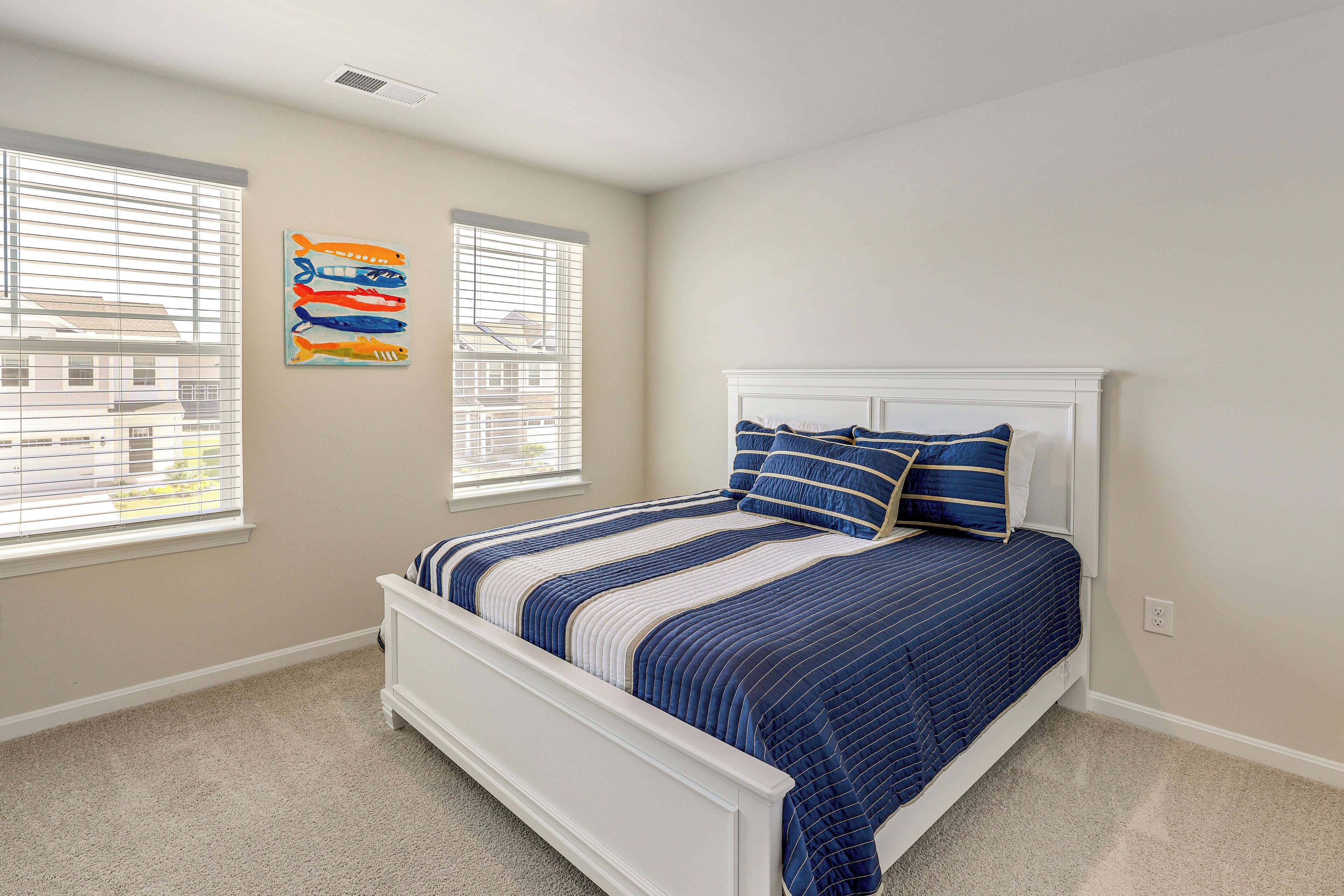 A bedroom with a white bed, blue and white striped bedding, and two windows with blinds.