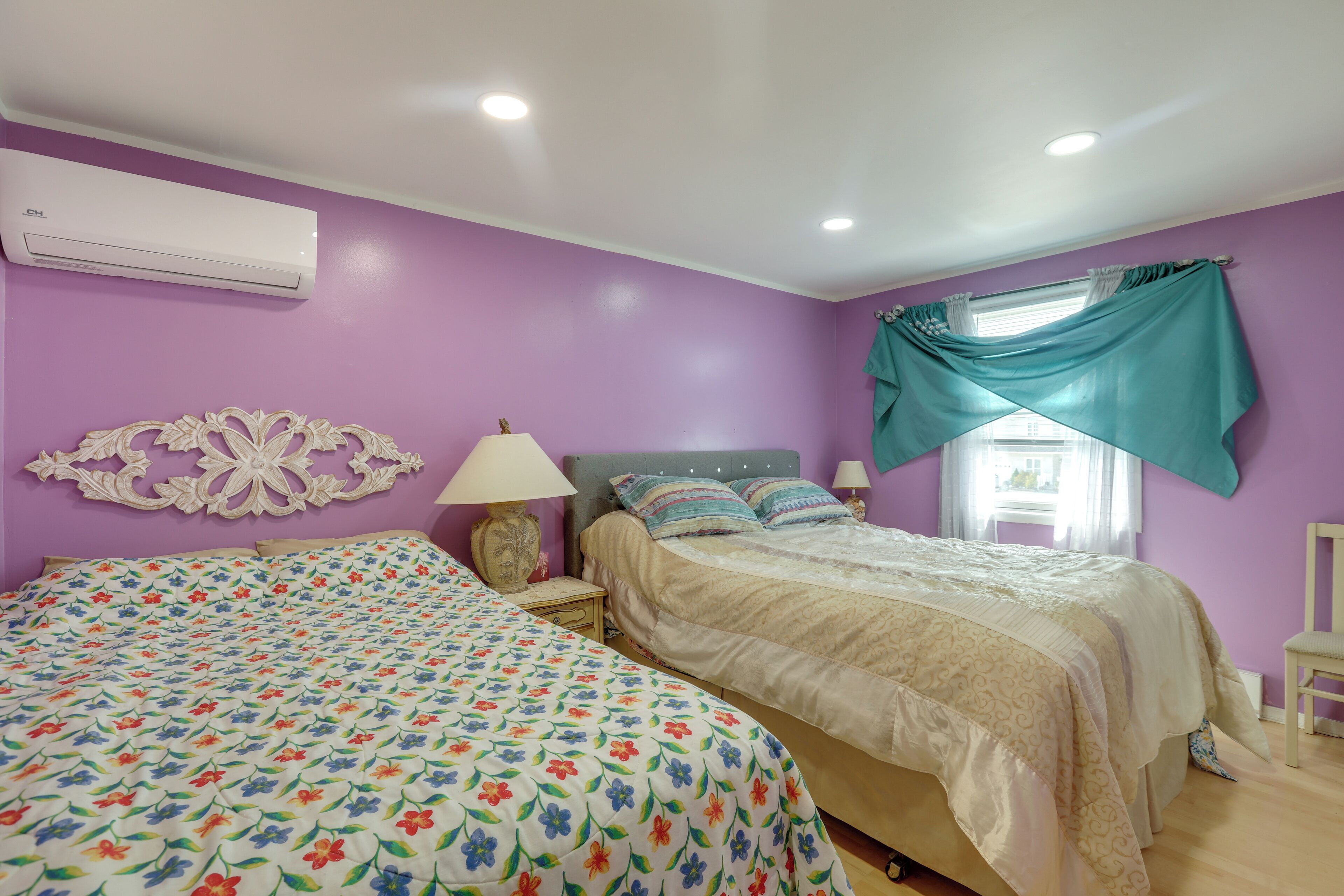 A bedroom with two beds, purple walls, a decorative wall piece, a window with curtains, and an air conditioning unit.