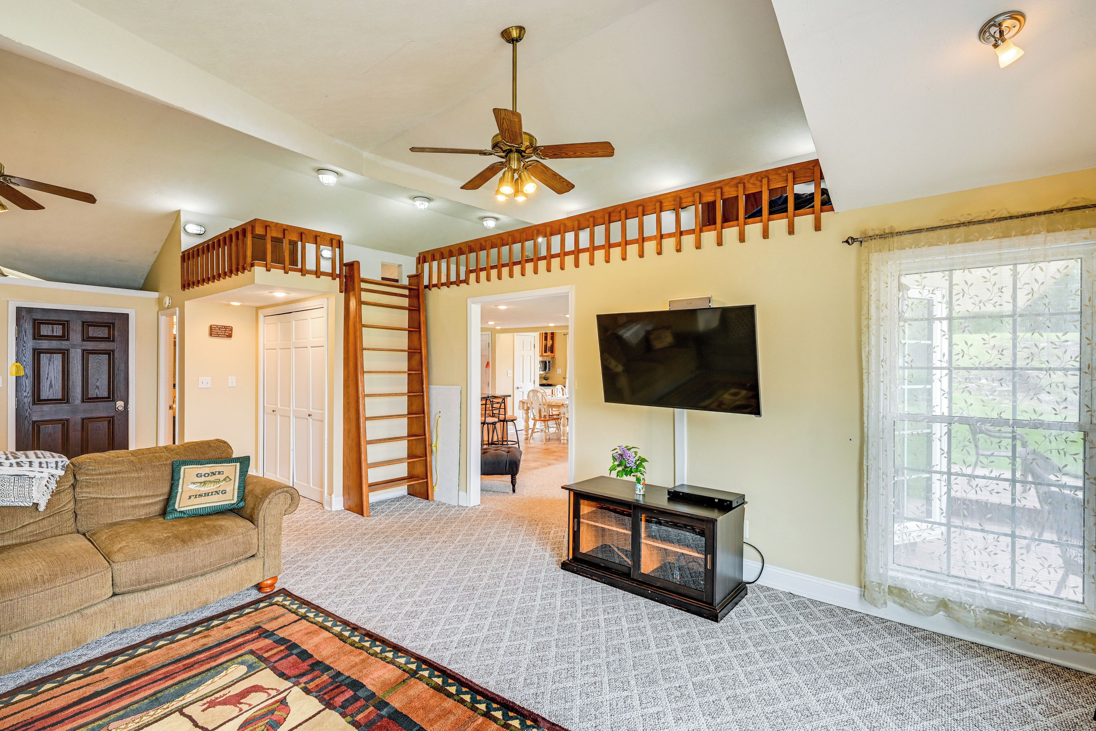 A spacious living room with a high ceiling, a ceiling fan, a large flat-screen TV, a wooden staircase, and a large window with a view.
