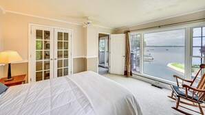 A bedroom with a large bed, a rocking chair, a nightstand, a lamp, and a view of the water through a large window. - Scenic Views: Spacious Webster Lake House! (North Webster)