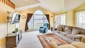 A spacious living room with a large window, a ceiling fan, a flat-screen TV, a blue recliner, a beige sofa, and a patterned rug. - Scenic Views: Spacious Webster Lake House! (North Webster)