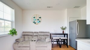 A modern living room with a grey sofa, a dining table with two chairs, a kitchen area with a refrigerator, and a wall-mounted decorative piece.