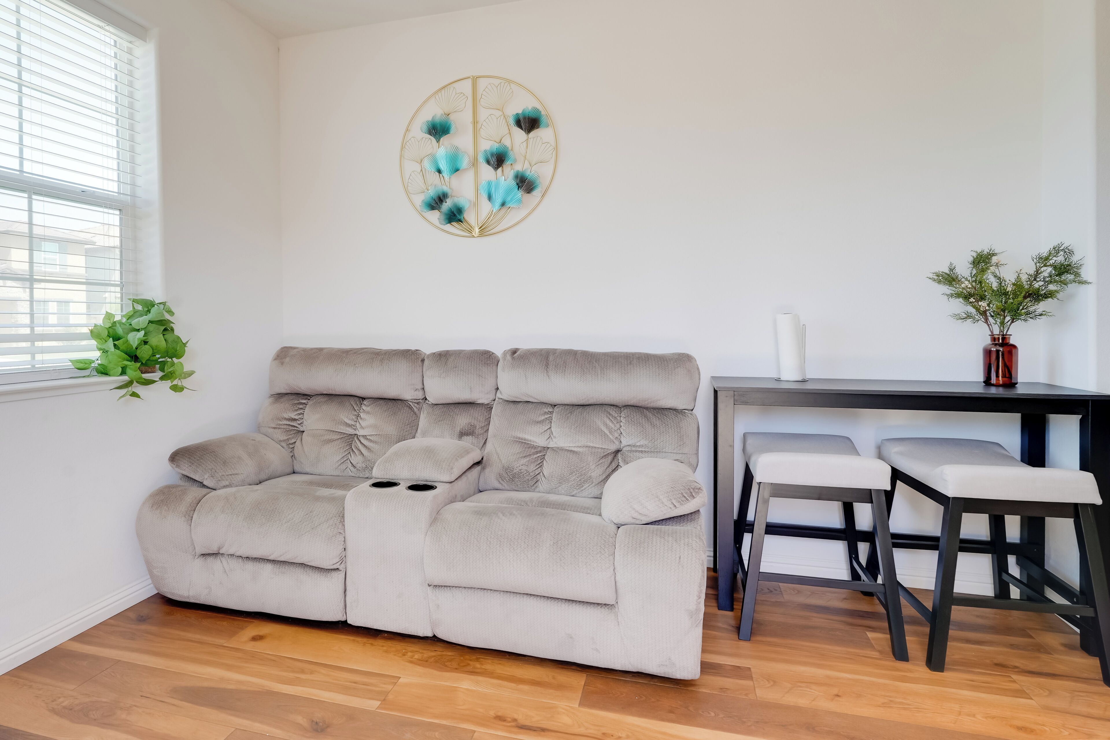 Una sala de estar moderna con un sofá beige, un comedor con una mesa pequeña y dos sillas, un elemento decorativo en la pared y una planta en maceta.