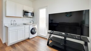 A compact kitchen with white cabinets, a microwave, a washing machine, and a large flat-screen TV on a black stand.