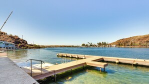 A waterfront property with a dock extending into clear water, a modern building with a balcony, and a hilly landscape in the background. - Game Room + Dock: Riverfront Escape in Parker! (Parker)