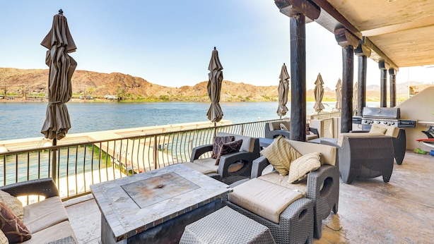 A covered outdoor seating area with wicker furniture and umbrellas, overlooking a body of water and hills. - Game Room + Dock: Riverfront Escape in Parker! (Parker)