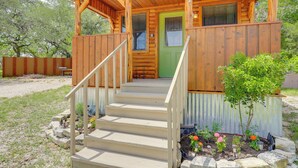 A wooden cabin with a green door, a covered porch, and a small garden with flowers. - Cozy Couple's Cabin Retreat in Concan! (Concan)