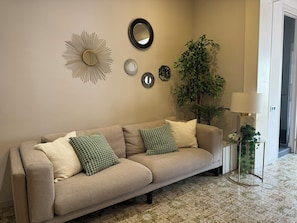 Una sala de estar con un sofá color beige, cojines estampados, un espejo y una planta en maceta.
