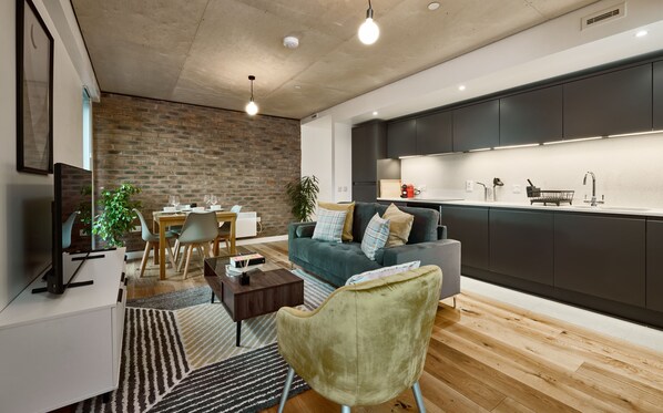 A modern living room with a dining area, a grey sofa, a green armchair, and a kitchen with dark cabinets and a white countertop.