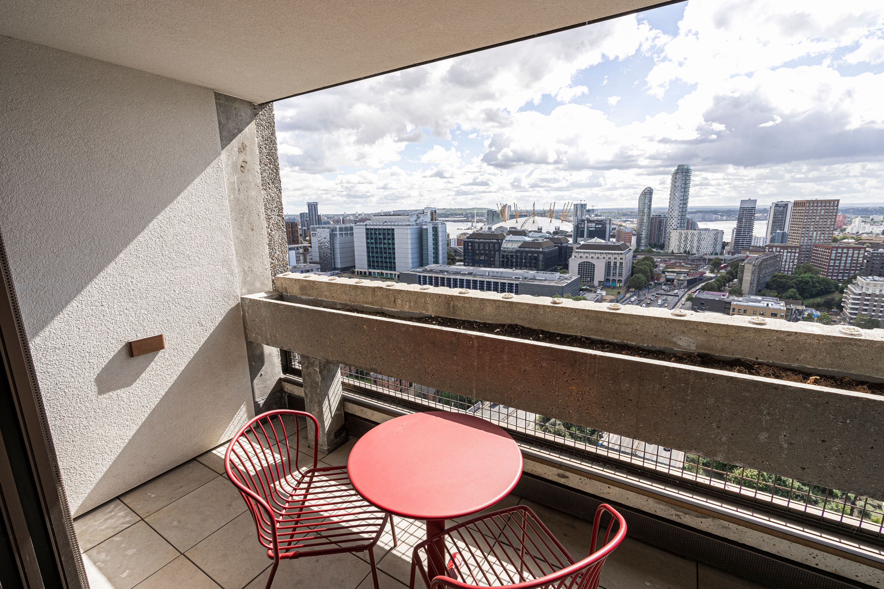 Un balcone con tavolo e sedie rosse, affacciato su una città con edifici moderni e cielo sereno.