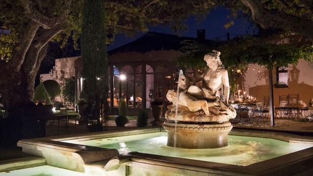 A nighttime outdoor fountain with a statue, surrounded by trees and a building with outdoor seating. - Charming Mas Provencal With Swimming Pool (Noves)
