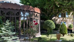 A conservatory with a glass roof, surrounded by well-maintained gardens and a traditional building in the background. - Charming Mas Provencal With Swimming Pool (Noves)