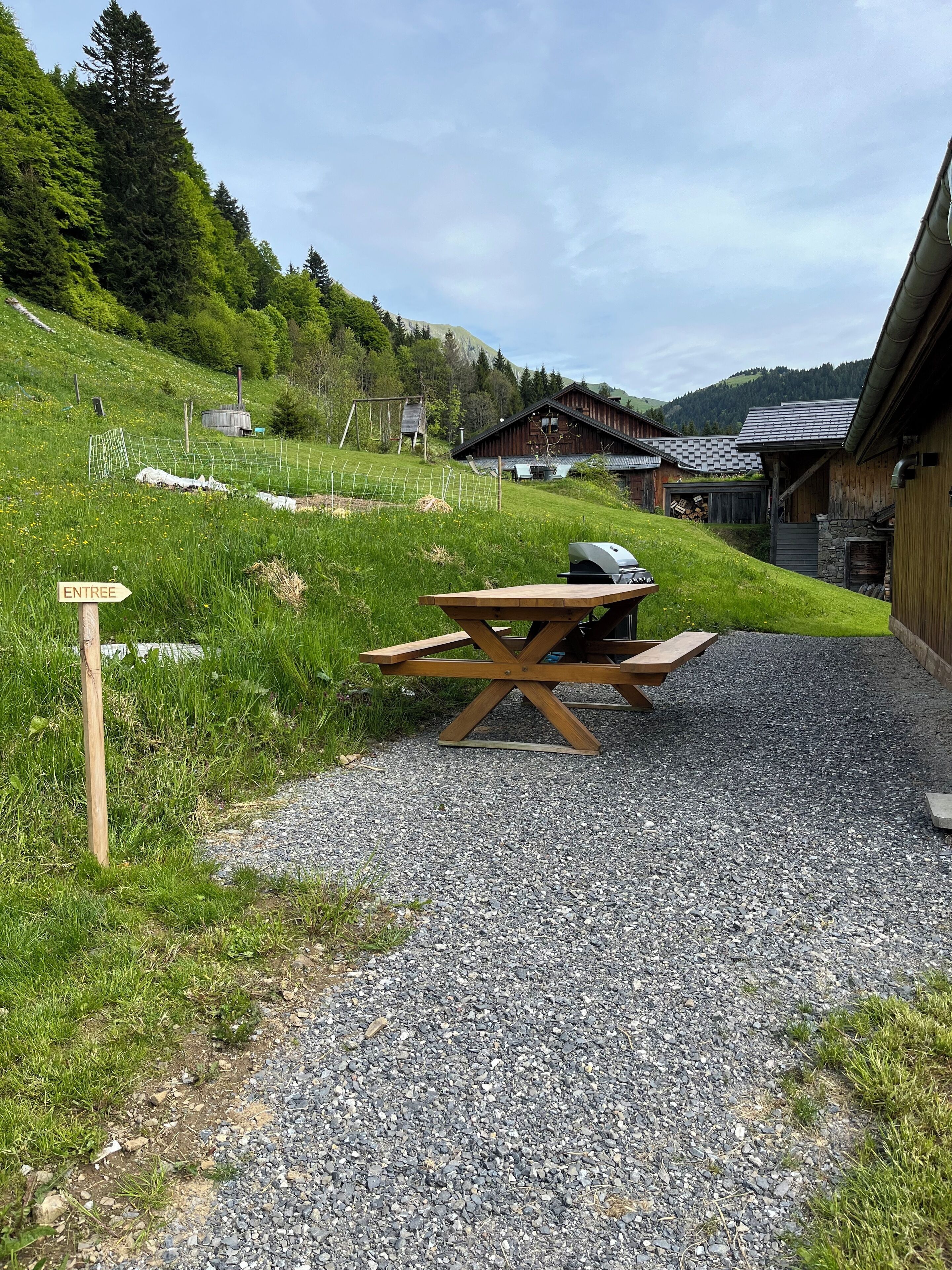 A picnic table with a grill in a grassy area with a gravel path, surrounded by wooden buildings and a sign pointing to 'ENTREE'.
