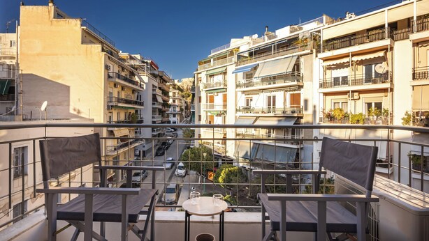 Un balcon avec deux chaises et une petite table, donnant sur une rue bordée de voitures garées et d’immeubles.