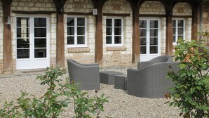 A building with stone walls and wooden beams, featuring a patio area with wicker furniture and a gravel ground. - Charming stone house 13 p, private garden & fireplace near Baie de Somme (Beauchamps)