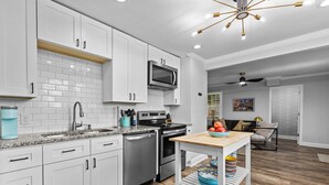A modern kitchen with white cabinets, a stainless steel stove, and a microwave. There's a wooden table with a bowl of fruit and a coffee maker on the counter.