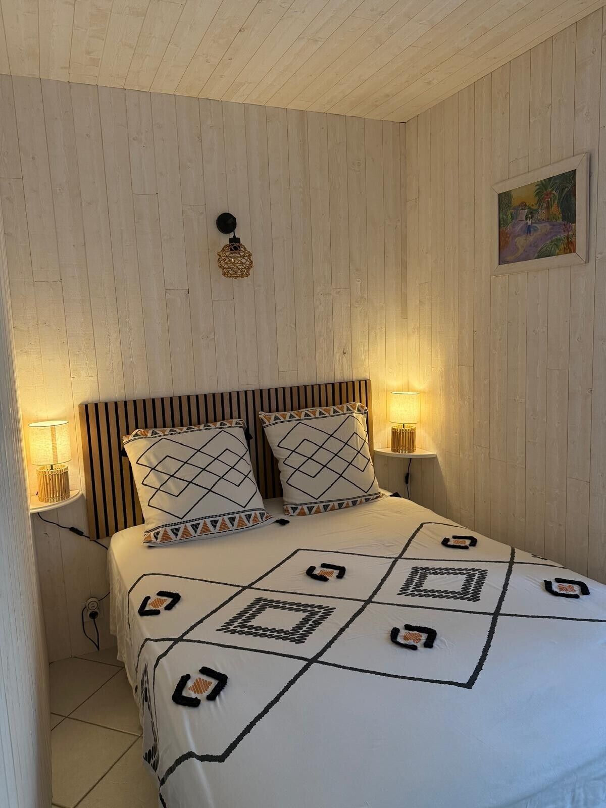 A bedroom with a wooden ceiling, a bed with patterned bedding, two bedside lamps, and a painting on the wall.