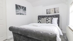 Un dormitorio con una cama tapizada en gris, ropa de cama blanca y un banco tapizado en gris. Hay dos obras de arte colgadas en la pared y un radiador blanco.
