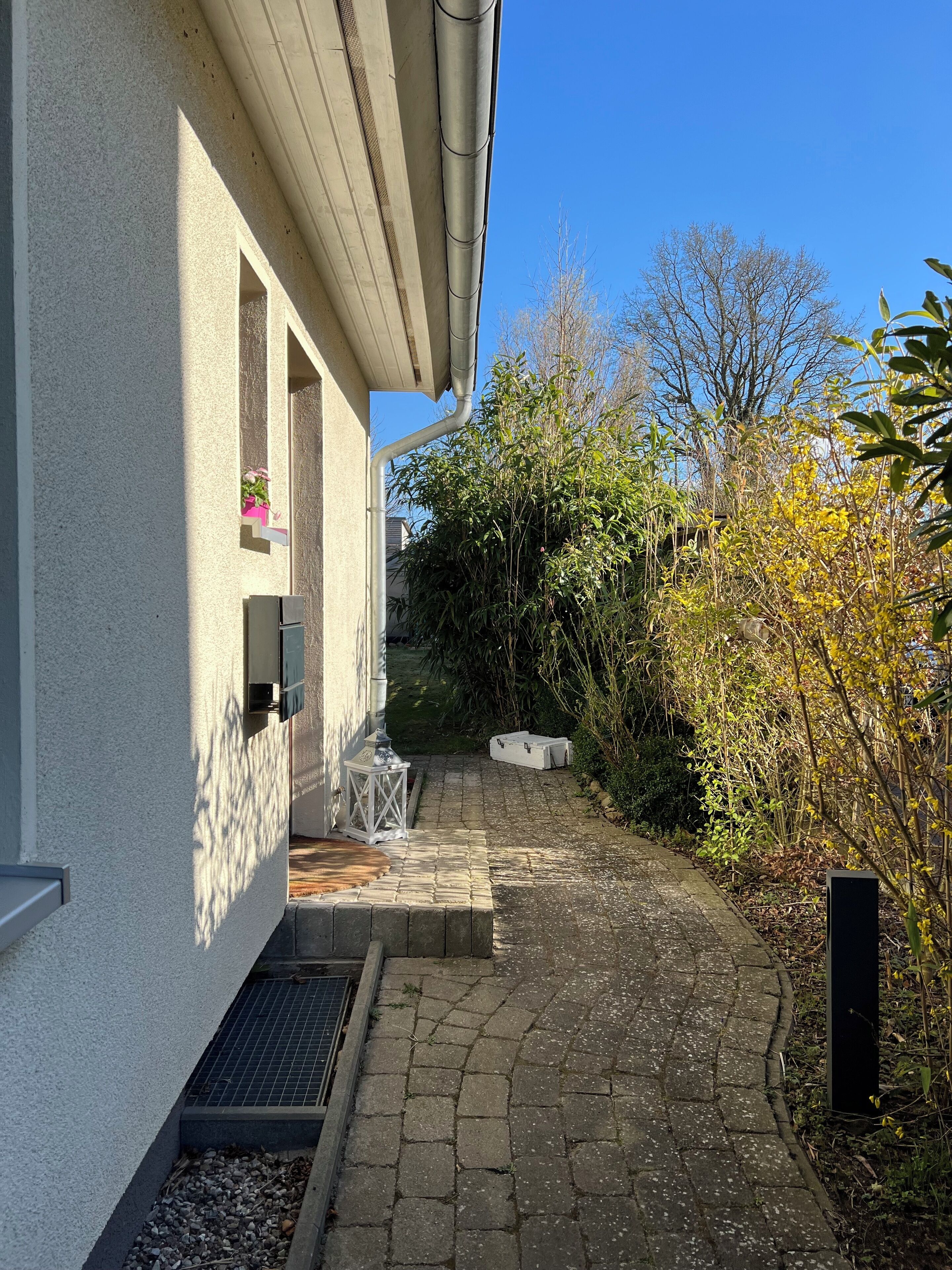 A paved pathway leading to a building with a white exterior and a small garden area with plants and a lantern.