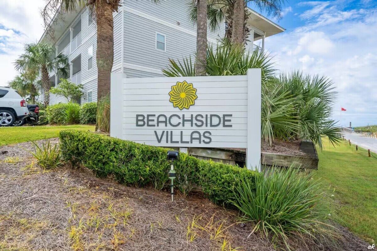 A sign for Beachside Villas with a yellow flower logo, surrounded by greenery and a white building in the background.