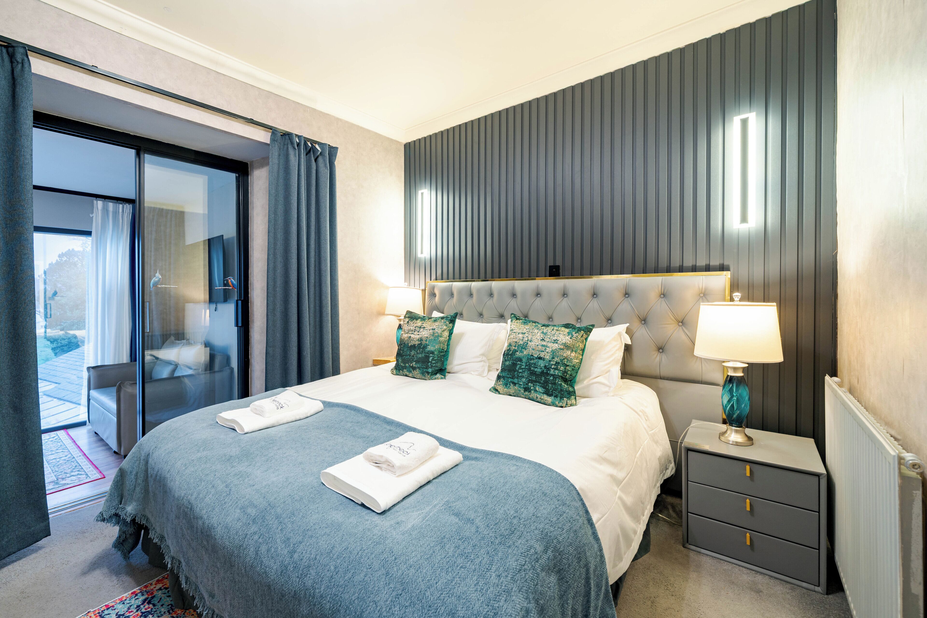 A hotel room with a large bed, a nightstand with a lamp, a sliding glass door to a balcony, and a view of the outdoors.