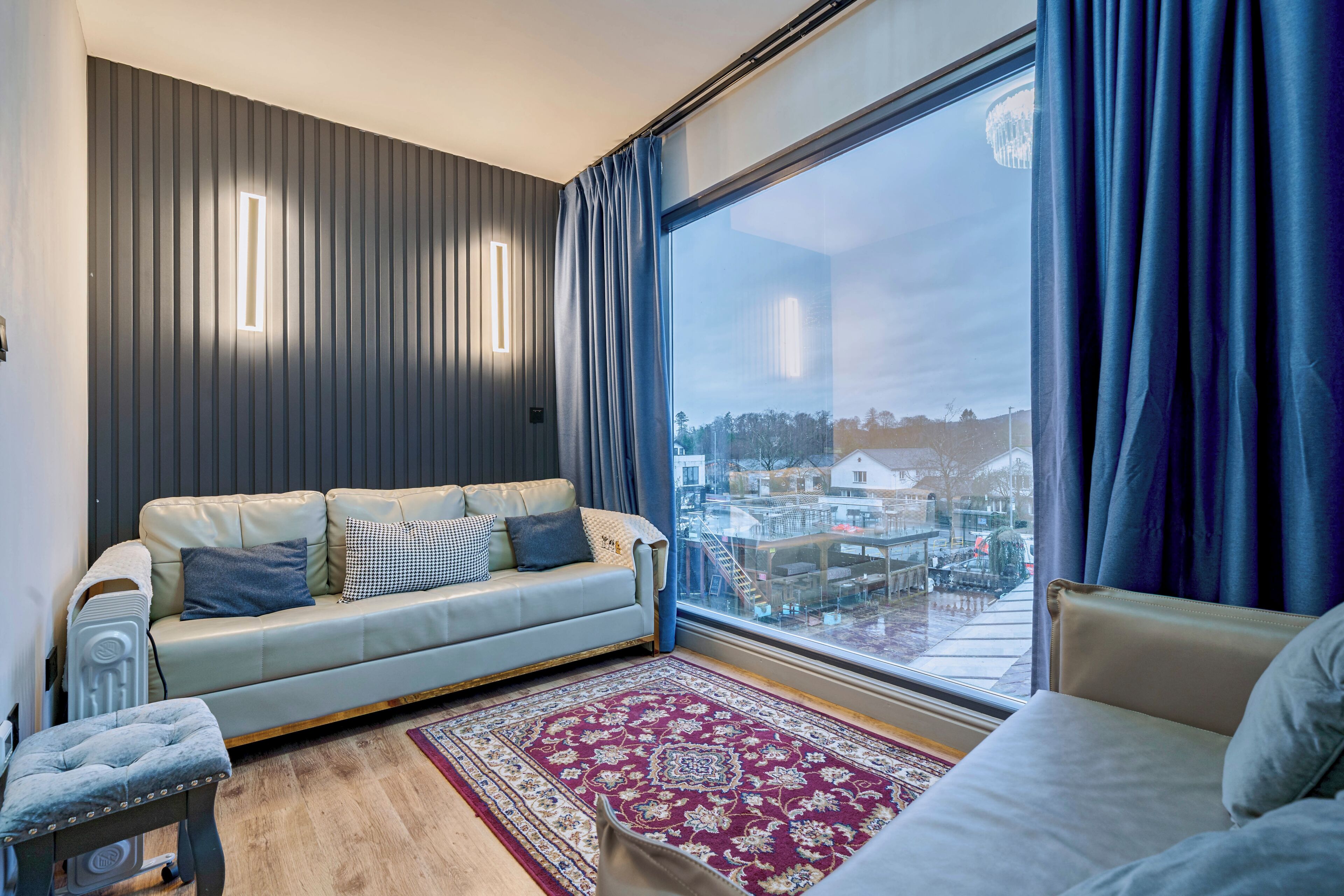 A modern living room with a sofa, a small table, a large window with curtains, and a view of buildings.