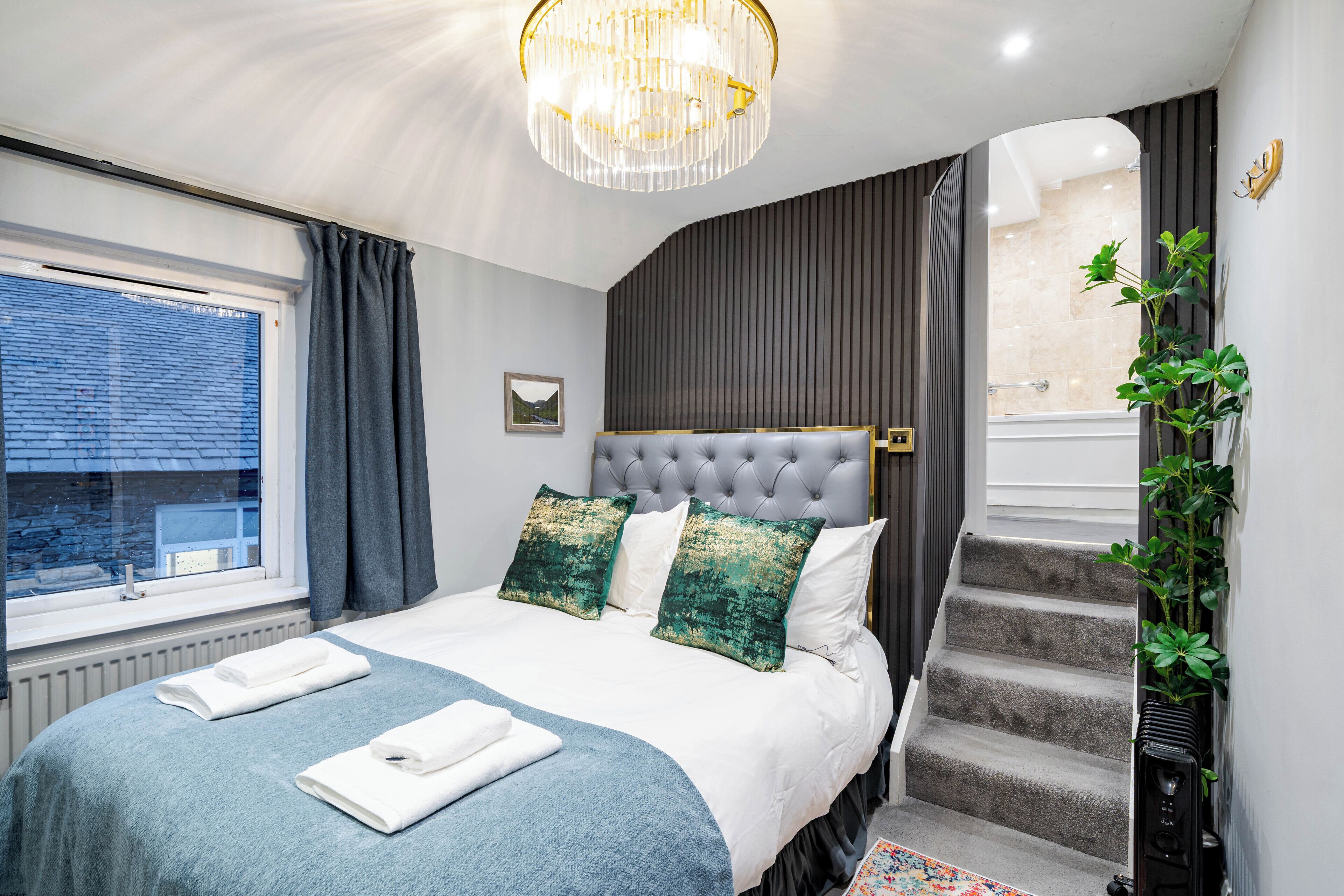 A bedroom with a large bed, a chandelier, a view through a window, and a staircase.