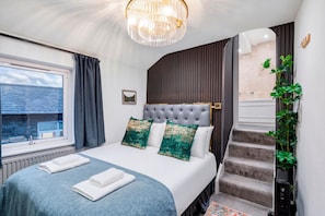 A bedroom with a large bed, a chandelier, a view through a window, and a staircase.