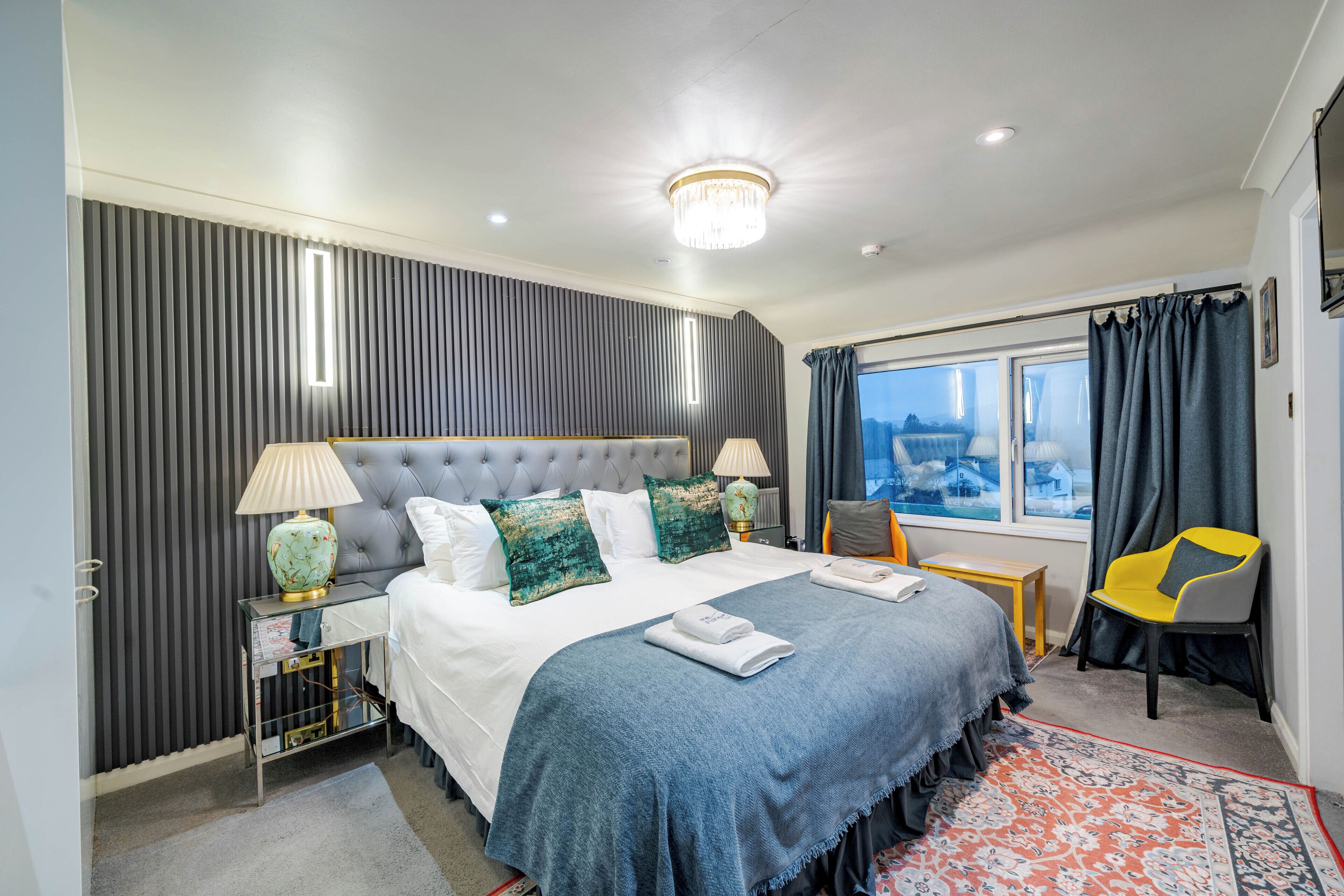 A hotel room with a large bed, a window with curtains, a chair, and a nightstand with a lamp.