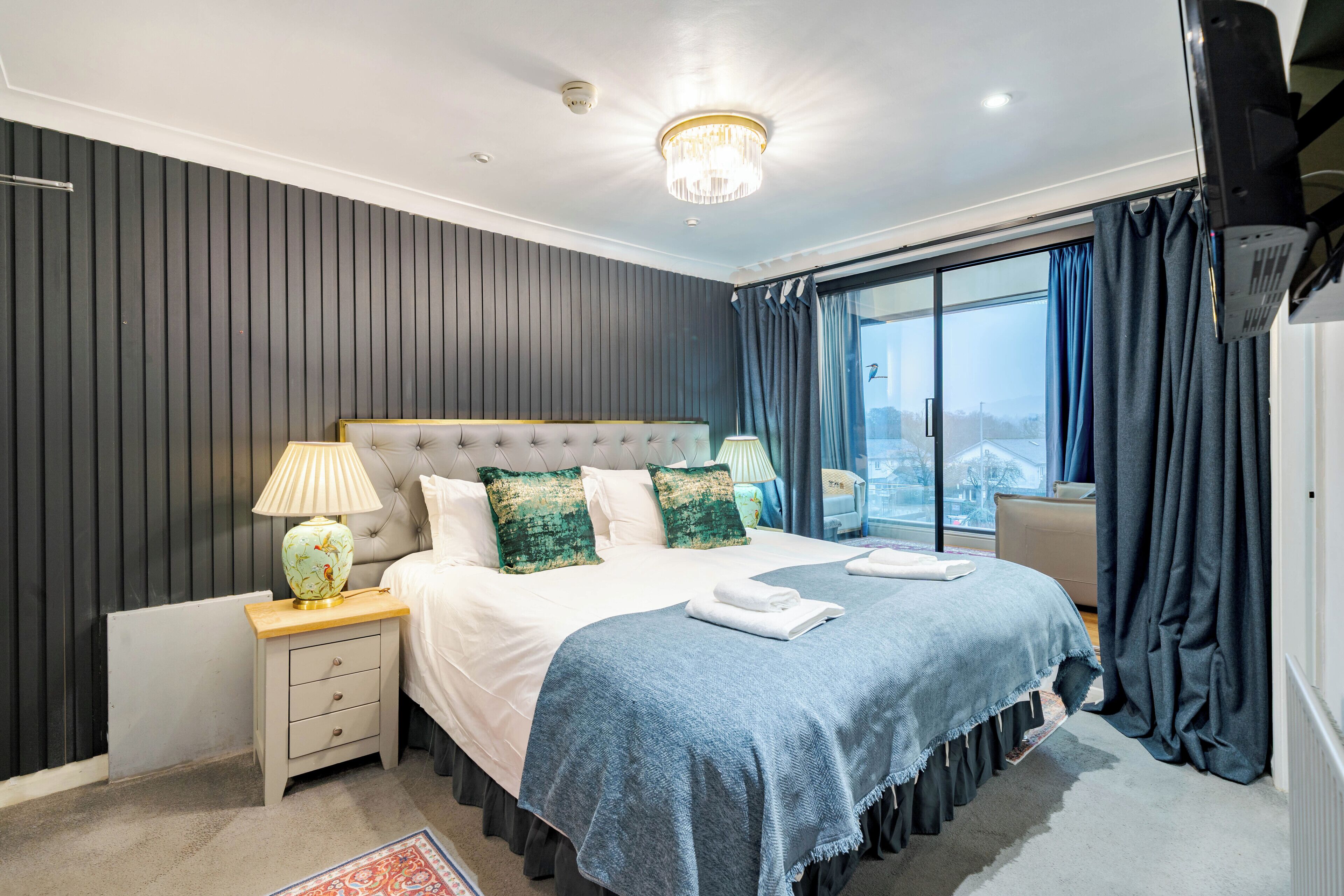 Une chambre à coucher moderne avec un grand lit, une table de chevet, une lampe et une vue sur l’extérieur.