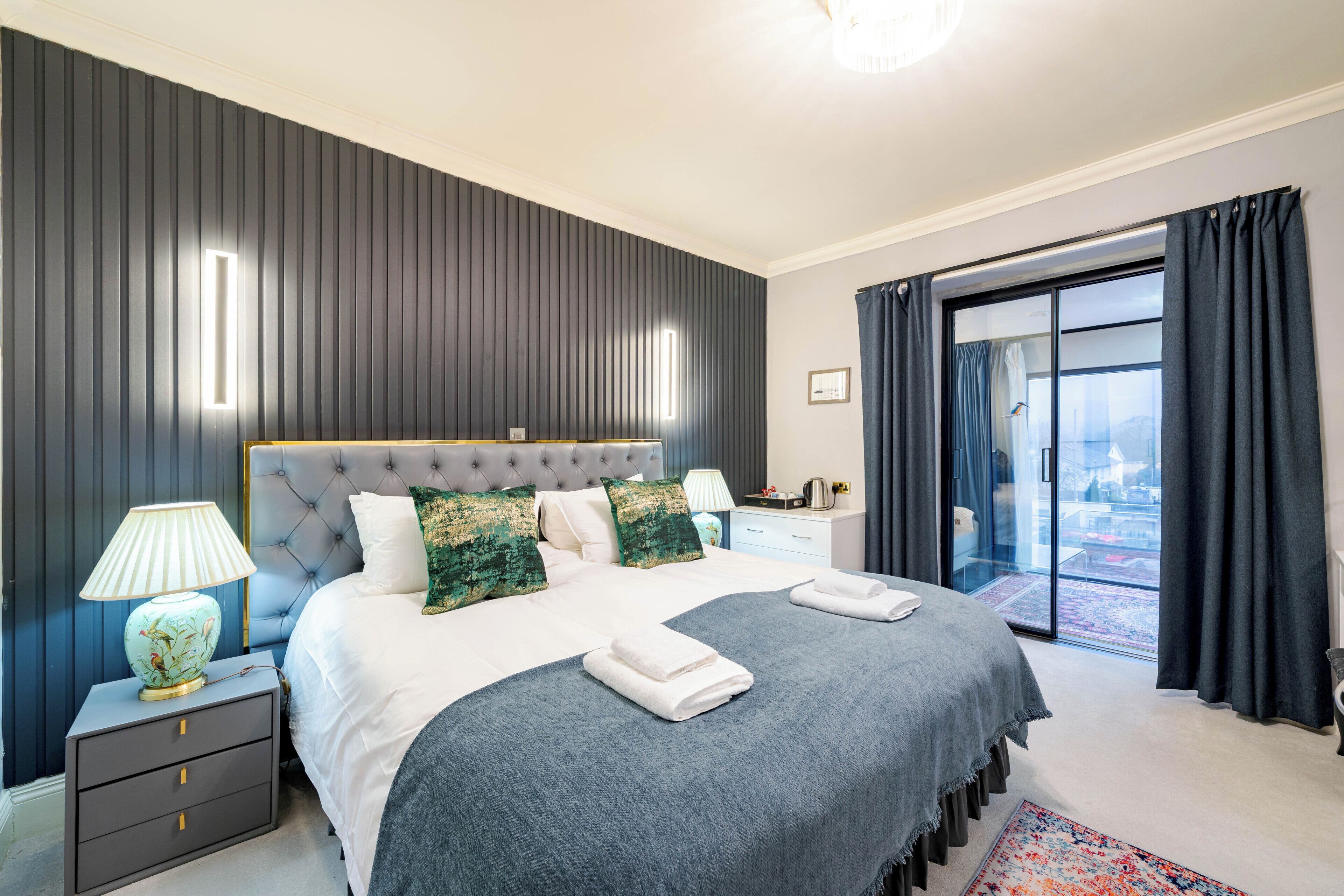 A bedroom with a large bed, a nightstand, a lamp, a window with curtains, and a view of the outside.