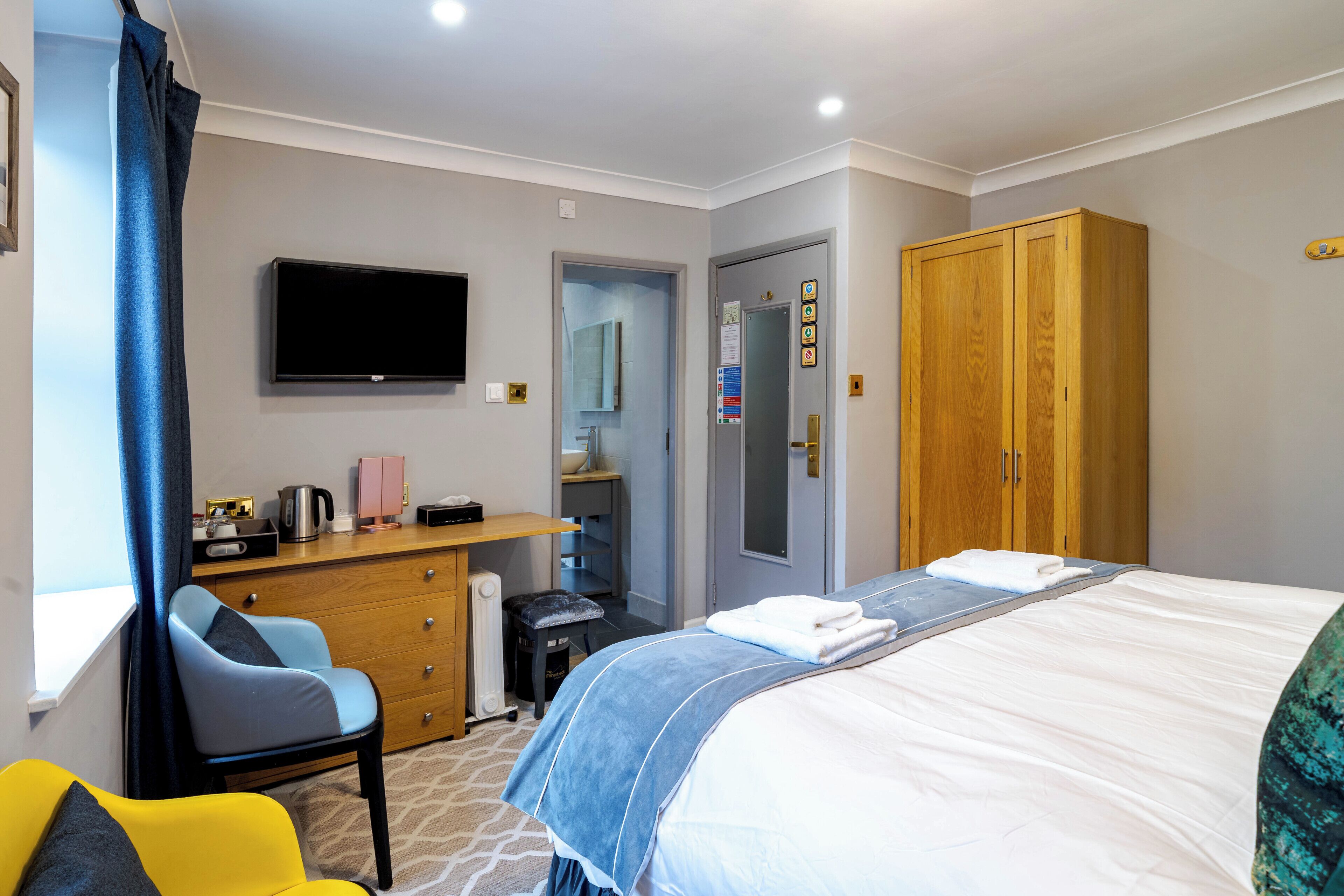 A hotel room with a bed, a TV, a wooden wardrobe, a desk with a chair, and a bathroom visible through an open door.