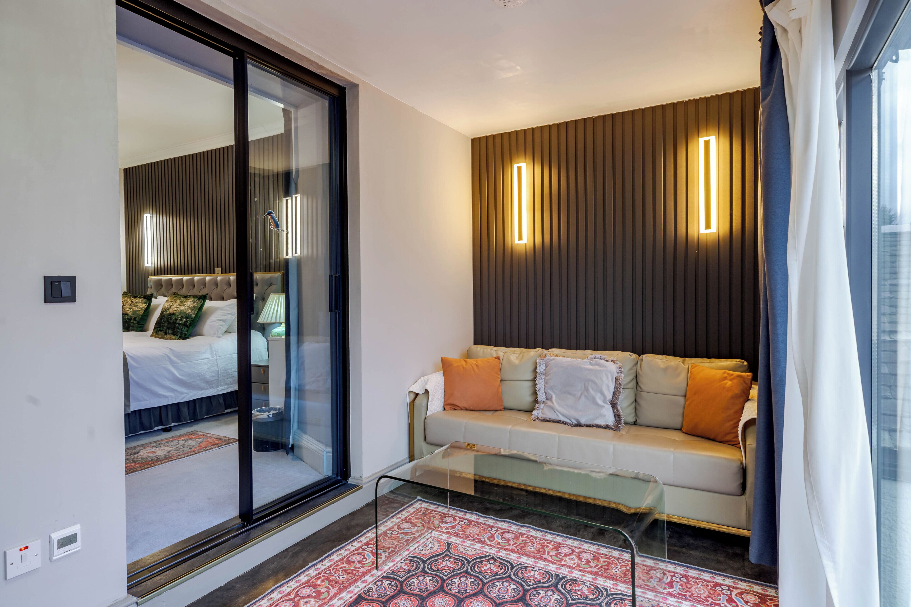 Un salon moderne doté d’une cloison en verre, d’un canapé avec des coussins, d’un tapis à motifs et d’un luminaire fixé au mur.