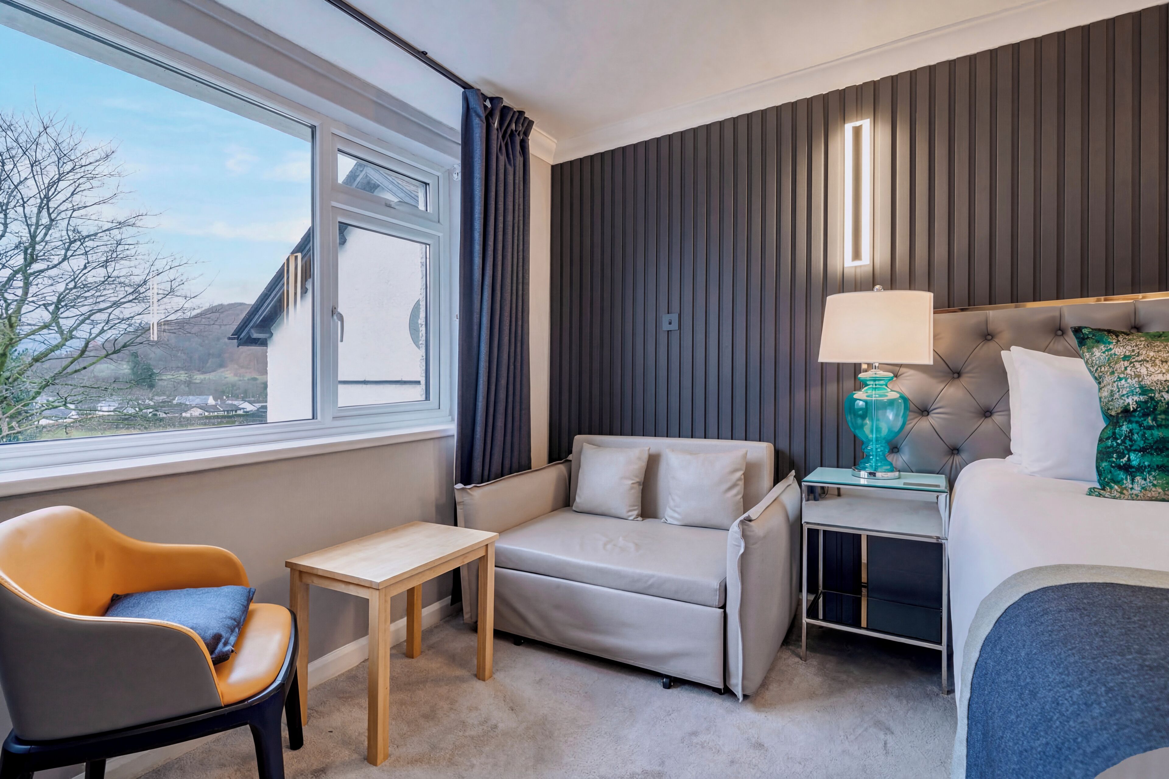 A hotel room with a sofa, a chair, a small table, a bedside lamp, and a bed.