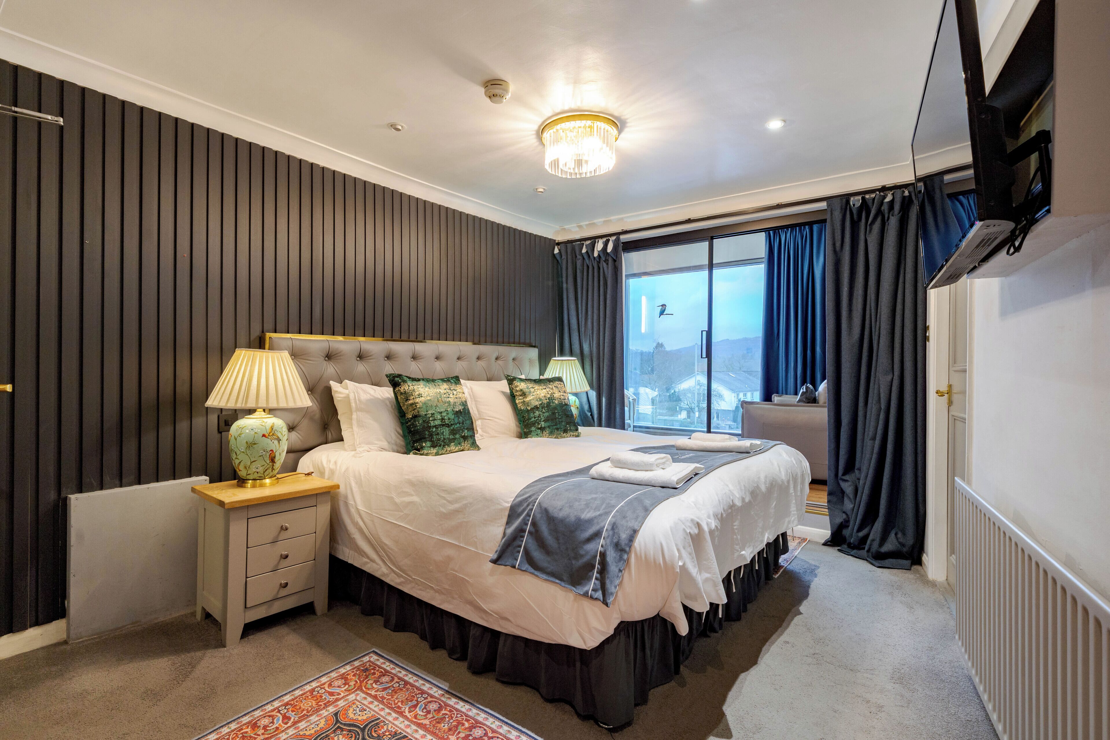 Une chambre avec un grand lit, une table de chevet, une lampe, un radiateur et une vue sur l’extérieur.
