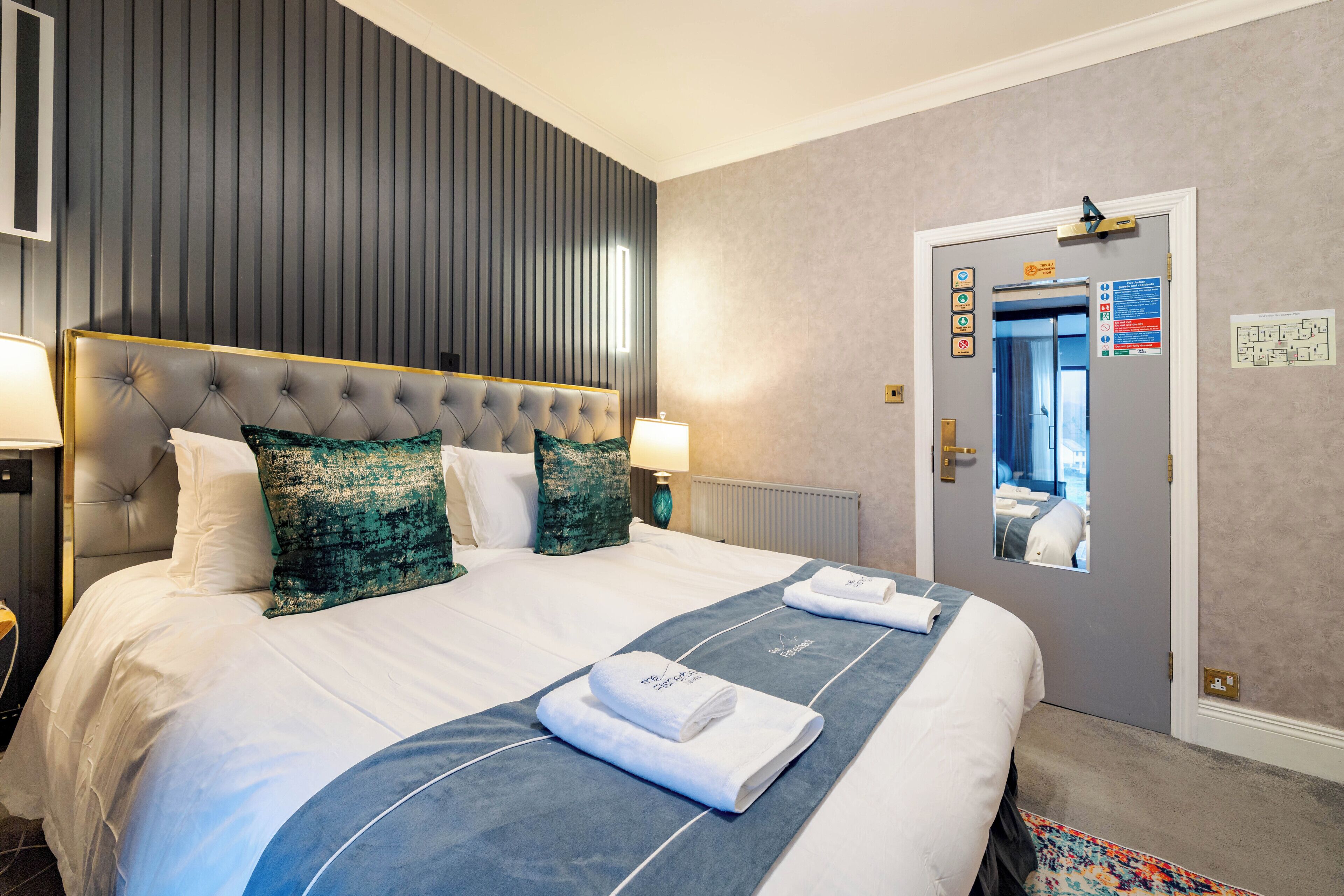 A hotel room with a large bed, a headboard, a mirror, and a door with an elevator sign.