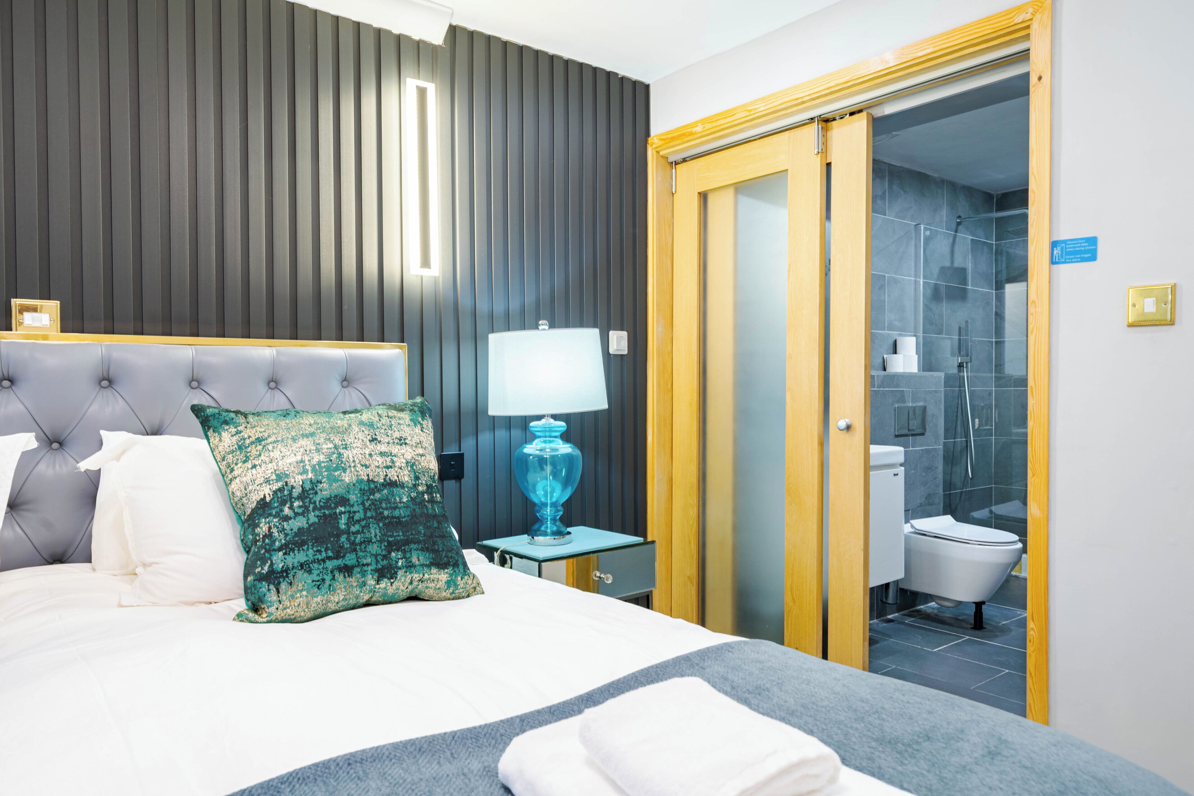 Ein Hotelzimmer mit Bett, Nachttisch mit Lampe, Badezimmer mit Dusche und Holztür.