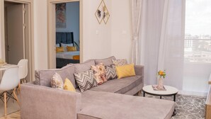 A modern living room with a grey sofa, a small round table, and a view of the city through sheer curtains. - Elite Residence  (Nairobi)